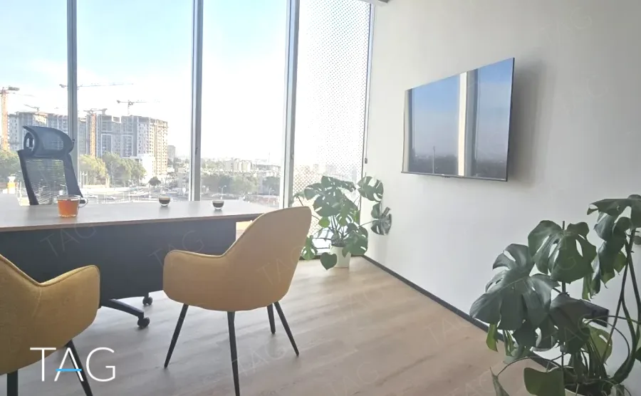 חדר עבודה בACRO BUSINESS CAMPUS עם שולחן, כורסאות, צמחייה ומסך על הקיר - תג נכסים
