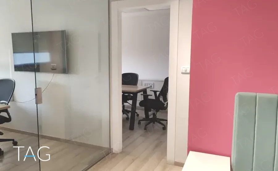 חדר עבודה קטן מאחורי מחיצת זכוכית במשרד להשכרה במגדל המאה, עם מסך תלוי על הקיר וקיר מג'נטה