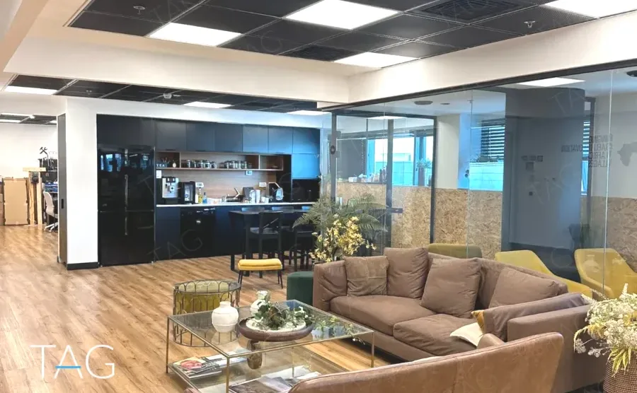חלל פתוח במשרד בבית אמפא הרצליה עם מטבחון כהה, ספות בהירות, פרקט עץ ומחיצות זכוכית - תג נכסים
