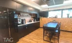 מטבחון כהה במשרד בבית אמפא הרצליה עם מכונת קפה, ארונות שחורים, שולחן בר גבוה וקיר עץ - תג נכסים