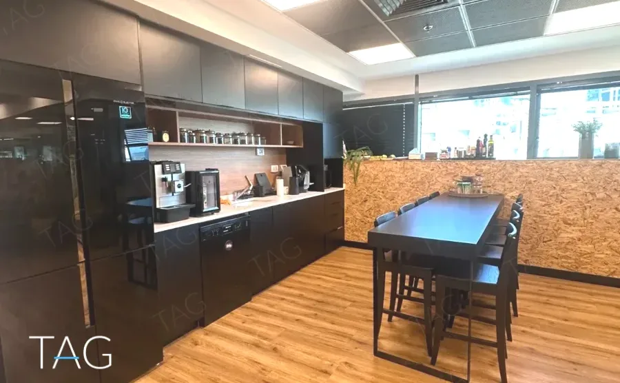 מטבחון כהה במשרד בבית אמפא הרצליה עם מכונת קפה, ארונות שחורים, שולחן בר גבוה וקיר עץ - תג נכסים