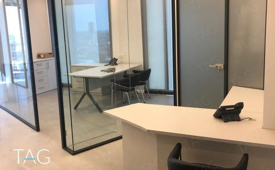 עמדת קבלה לבנה עם כיסא, מול חדר זכוכית ודלת כהה עם ידית בגוון פליז