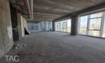חלל משרדי פתוח במצב מעטפת עם תקרת בטון חשופה, עמודי בטון וחלונות זכוכית גבוהים לנוף עירוני, תמונה להמחשה