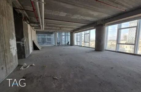 חלל משרדי פתוח במצב מעטפת עם תקרת בטון חשופה, עמודי בטון וחלונות זכוכית גבוהים לנוף עירוני, תמונה להמחשה