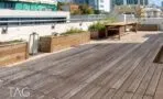 גג מרוצף בדק עץ עם ארגזי עץ מלאי צמחים ושולחן ארוך, מול בניינים בתל אביב