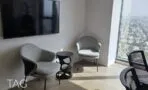 פינת ישיבה עם שתי כורסאות אפורות ושולחן צד מתכת לצד מסך קיר וחלון גבוה לנוף העיר, מגדלי אלון