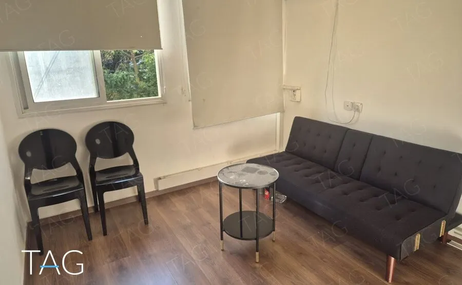 פינת המתנה עם ספה שחורה, שולחן צד עגול ושני כיסאות, מתחת לחלון עם וילון גלילה