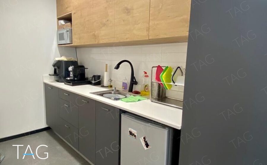 מטבחון במשרד להשכרה ברמת גן: ארונות עליונים בגוון עץ, ארונות תחתונים אפורים, משטח לבן, כיור ומקרר קטן