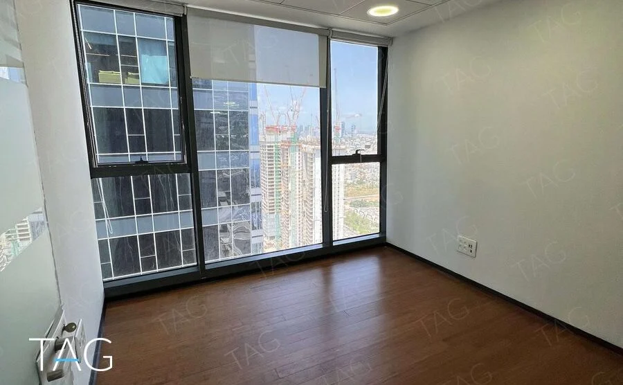 חדר משרד קטן עם חלון גבוה, תריס לבן ופרקט חום כהה, ובחוץ בנייני מגדלים ועגורני בנייה