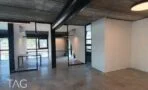 אולם משרדי עם מחיצות זכוכית במסגרות ברזל שחורות, קירות לבנים, רצפת בטון מוחלקת וצינור מיזוג שחור