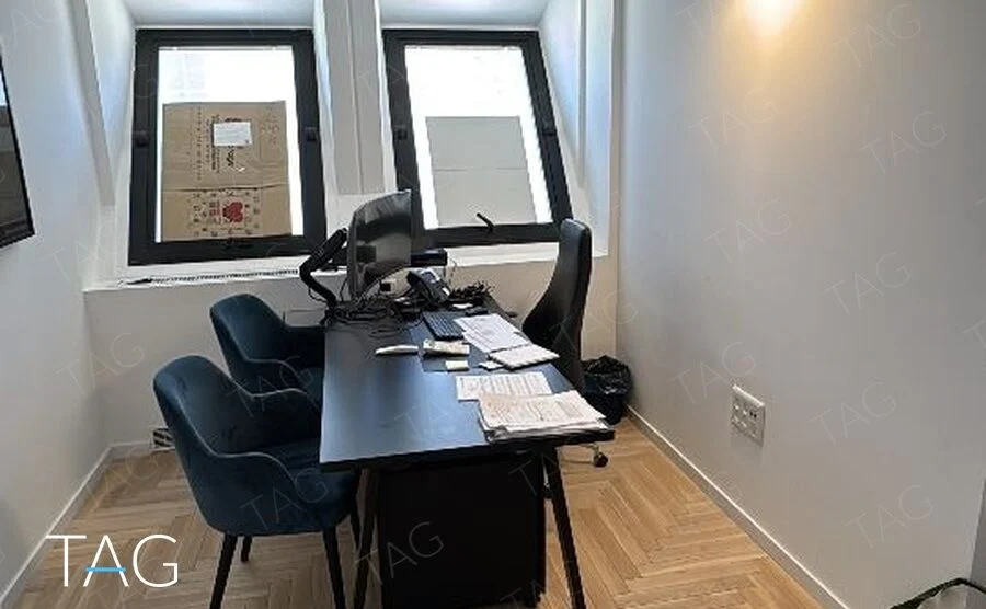 חדר עבודה צר עם שולחן שחור וכיסאות כחולים מתחת לשני חלונות משופעים ופרקט פישבון בבית הטקסטיל