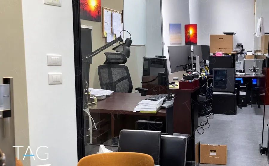 חדר מנהל עם שולחן כהה וכיסא ארגונומי, מבט לאזור עבודה עם מחשבים