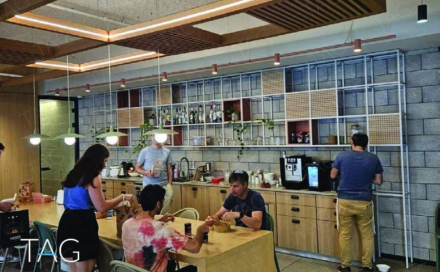 מטבחון משותף עם שולחן עץ גבוה, ארונות עץ בהירים ומדפי מתכת לבנים על קיר בלוקים